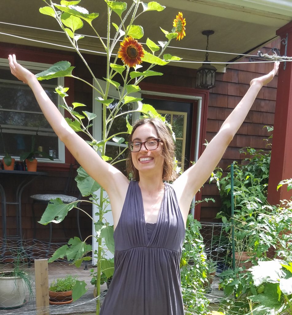 Photograph of Kim Adams with her arms in the air in triumph in front of a homegrown sunflower