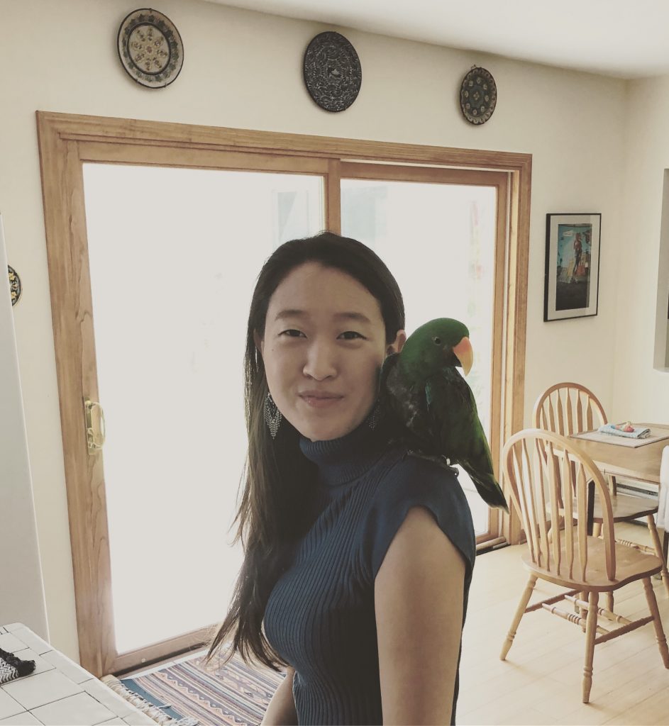 Color picture of Asian woman in a dark shirt with a parrot on her shoulder.