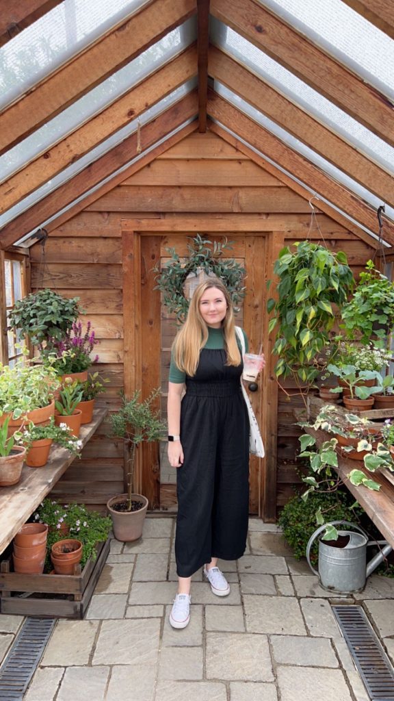 Picture of blonde Caucasian woman dressed in black and green with a bag over her shoulder standing in a greenhouse.