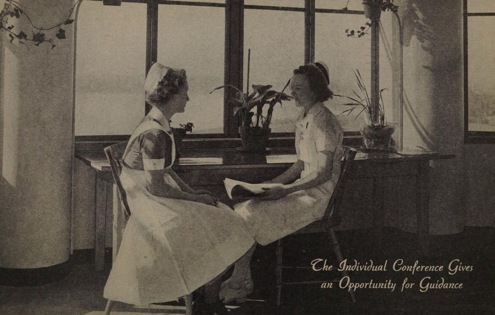Black and white photograph of two women in nursing attire sitting facing each other at a narrow table. Text on the photograph reads: "The individual conference gives an opportunity for guidance."